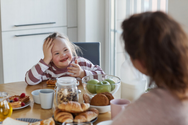 Kuvassa iloinen tyttö, jolla downin syndrooma, syömässä aamupalaa ja juttelemassa äidilleen.